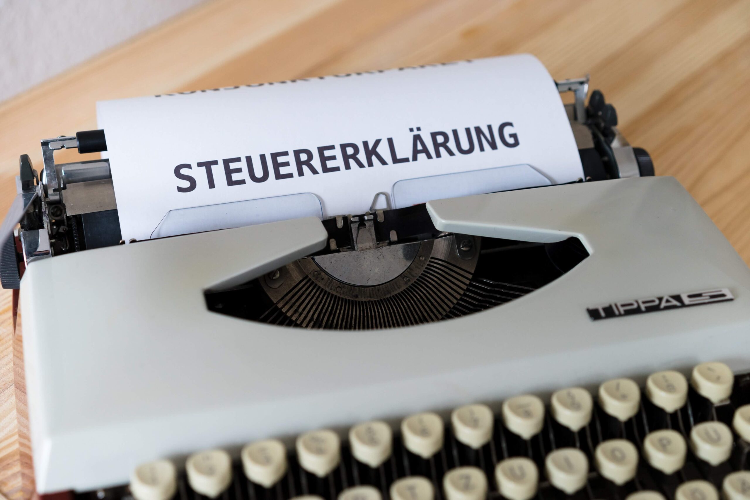 Schreibmaschine mit eingelegtem Papier, auf dem das Wort „STEUERERKLÄRUNG“ gedruckt ist, auf Holzschreibtisch.